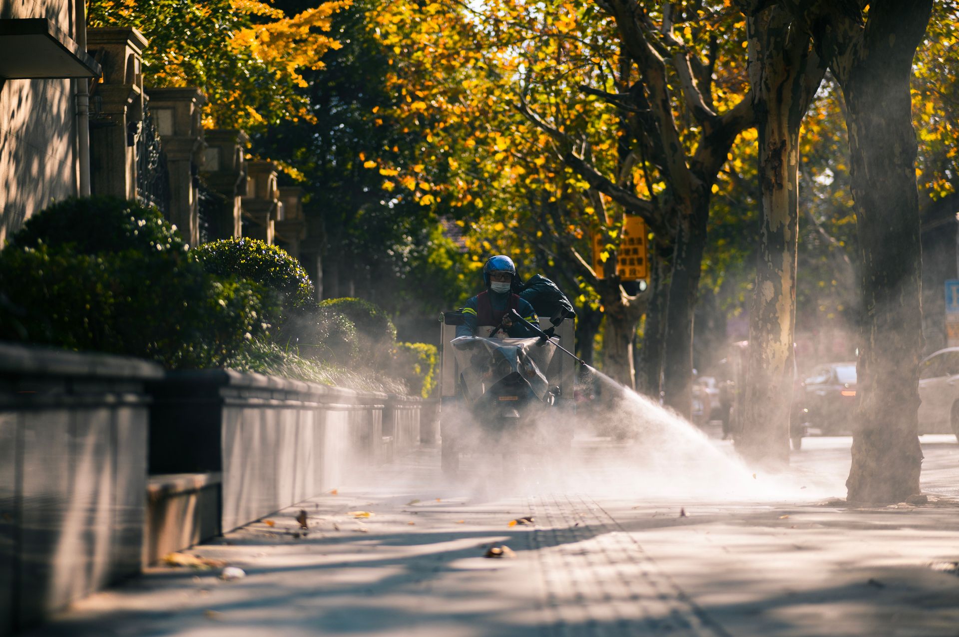 A city worker sprays a sidewalk with water on a sunny day, surrounded by lush trees and buildings.